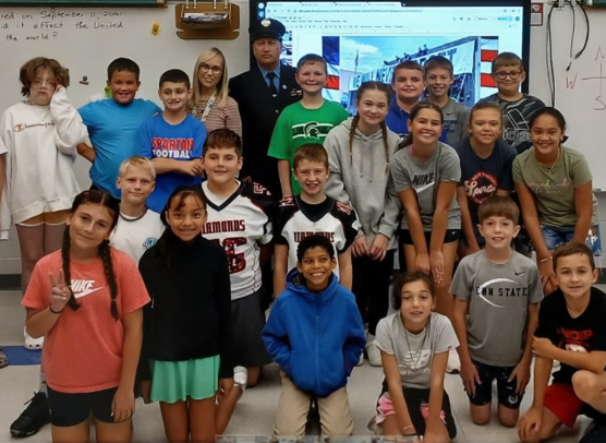 Dennis Prosick in FDNY dress uniform visiting an elementary school classroom as a guest speaker, posing with students and their teacher.